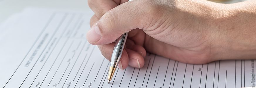 Eine Hand hält einen silbernen Kugelschreiber und füllt ein Formular aus. Das Formular enthält mehrere leere Felder für persönliche Informationen.