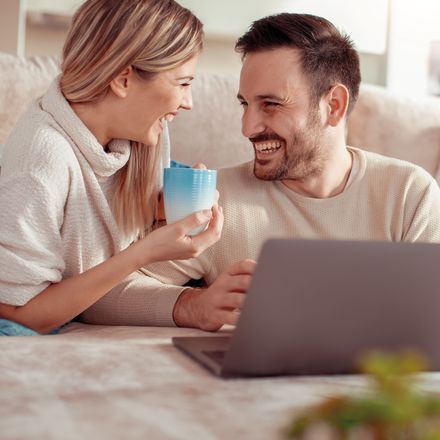 Ein Mann und eine Frau sitzen auf einem Sofa. Die Frau hält eine Tasse in der Hand und scheint mit dem Mann zu sprechen, während der Mann einen Laptop vor sich hat. Beide tragen helle Pullover und wirken entspannt. Im Vordergrund ist ein unscharfes grünes Objekt zu sehen, möglicherweise eine Pflanze.