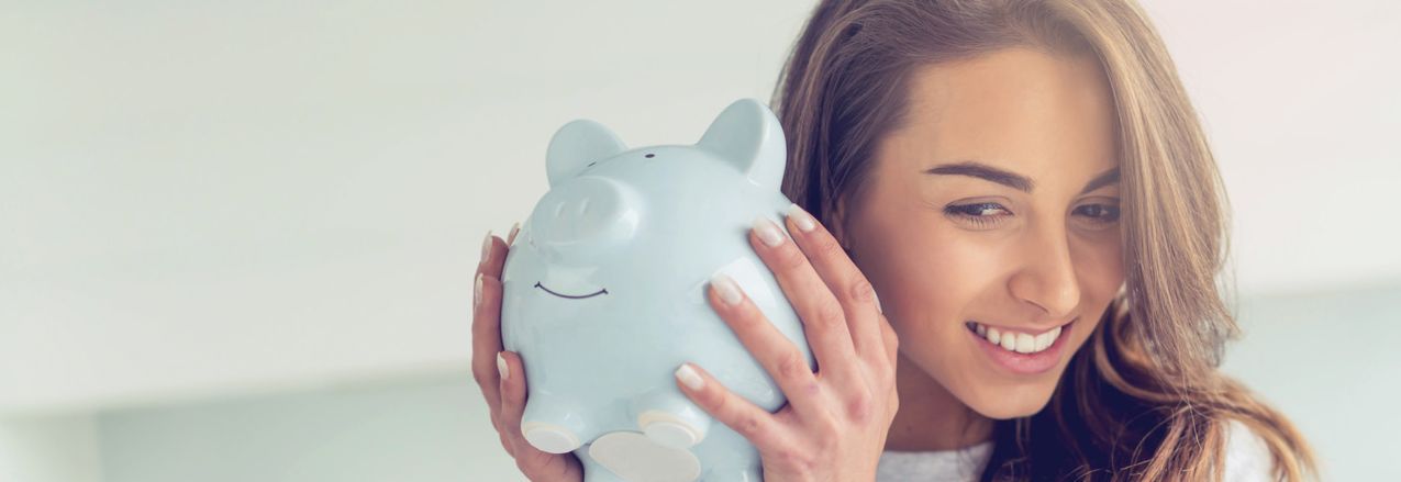 Eine Frau hält ein hellblaues Sparschwein mit beiden Händen neben ihrem Gesicht. Das Sparschwein hat ein lächelndes Gesicht und die Frau hat lange, braune Haare. Die Frau lächelt.