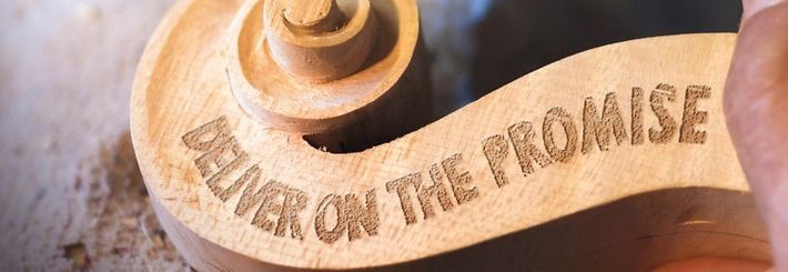 Eine Hand hält ein hölzernes Werkstück, auf dem die Worte "DELIVER ON THE PROMISE" eingraviert sind. Eine andere Hand bearbeitet das Holz mit einem Werkzeug. Das Bild zeigt eine handwerkliche Tätigkeit, bei der Holz geschnitzt oder graviert wird.