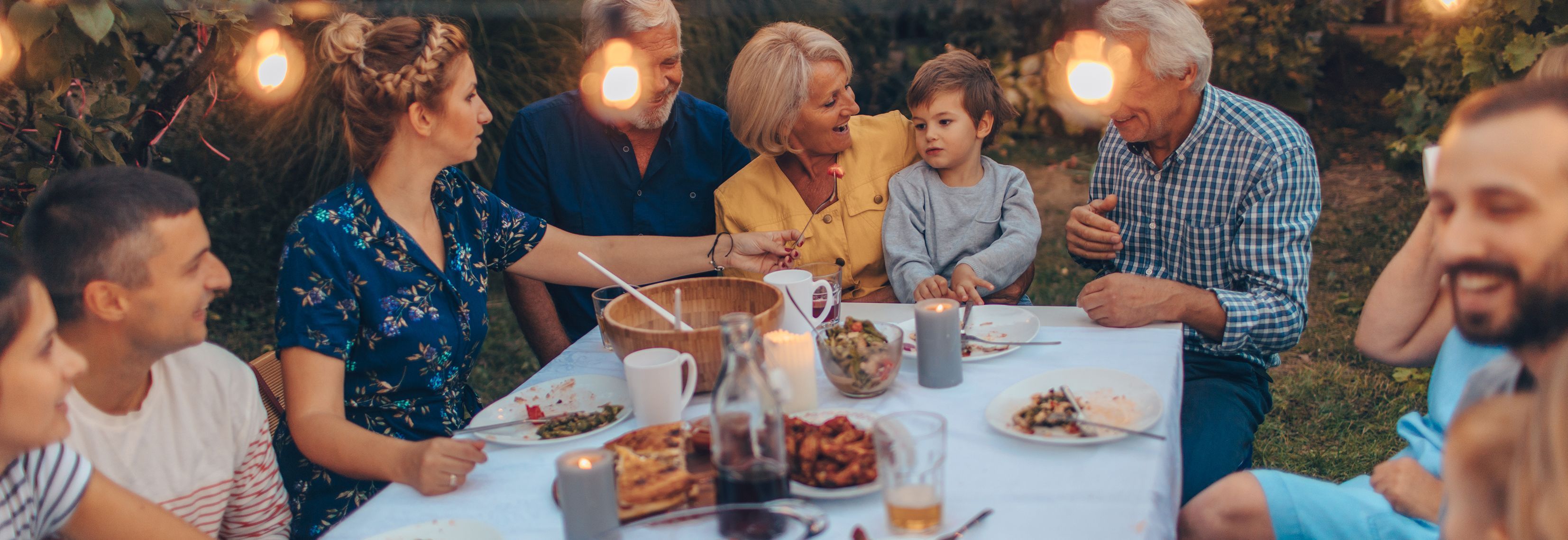 Eine Gruppe von Menschen sitzt an einem Tisch im Freien und isst zusammen. Der Tisch ist mit verschiedenen Speisen und Getränken gedeckt. Im Hintergrund sind Lichterketten zu sehen, die eine gemütliche Atmosphäre schaffen.