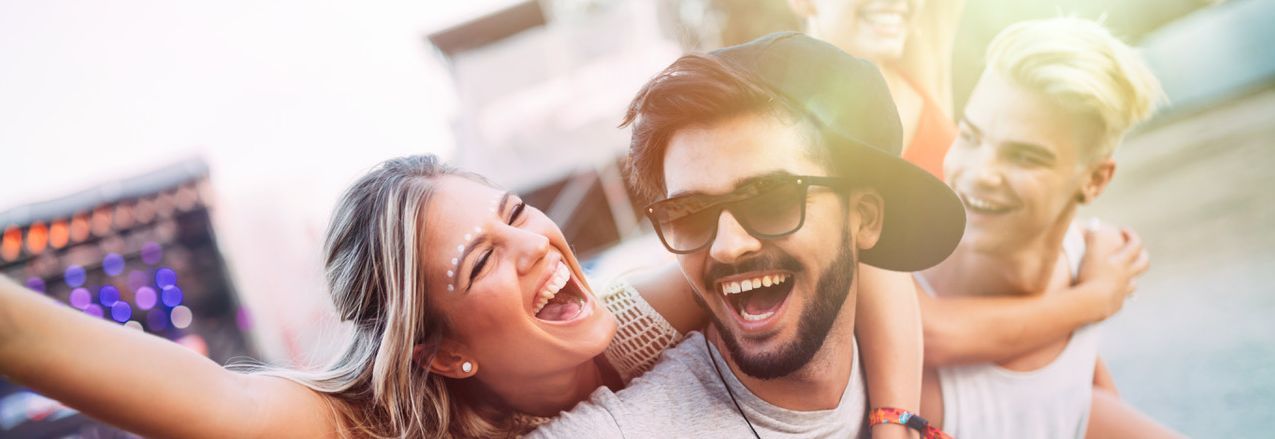 Das Bild zeigt eine Gruppe von Menschen, die zusammen Spaß haben. Im Vordergrund sind drei Personen zu sehen die am Lachen sind. Eine Person trägt eine Kappe und hat den Arm um eine andere Person gelegt. Im Hintergrund ist ein verschwommenes Stadtbild oder eine Veranstaltung zu erkennen.