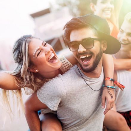 Das Bild zeigt eine Gruppe von Menschen, die zusammen Spaß haben. Im Vordergrund sind drei Personen zu sehen die am Lachen sind. Eine Person trägt eine Kappe und hat den Arm um eine andere Person gelegt. Im Hintergrund ist ein verschwommenes Stadtbild oder eine Veranstaltung zu erkennen.