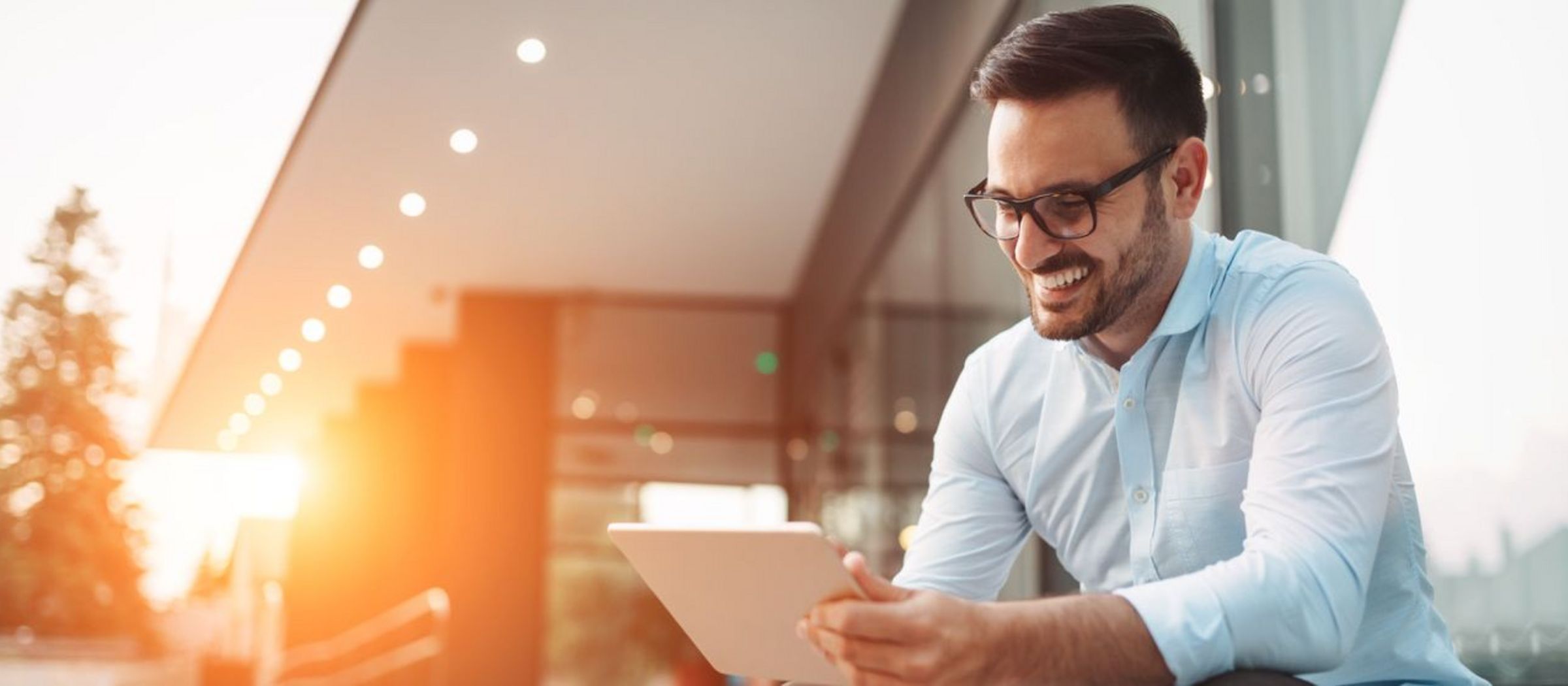 Ein Mann sitzt draußen und hält ein Tablet in den Händen. Er trägt ein weißes Hemd und eine Brille. Im Hintergrund ist ein modernes Gebäude mit Glasfassade zu sehen, das von der untergehenden Sonne beleuchtet wird.