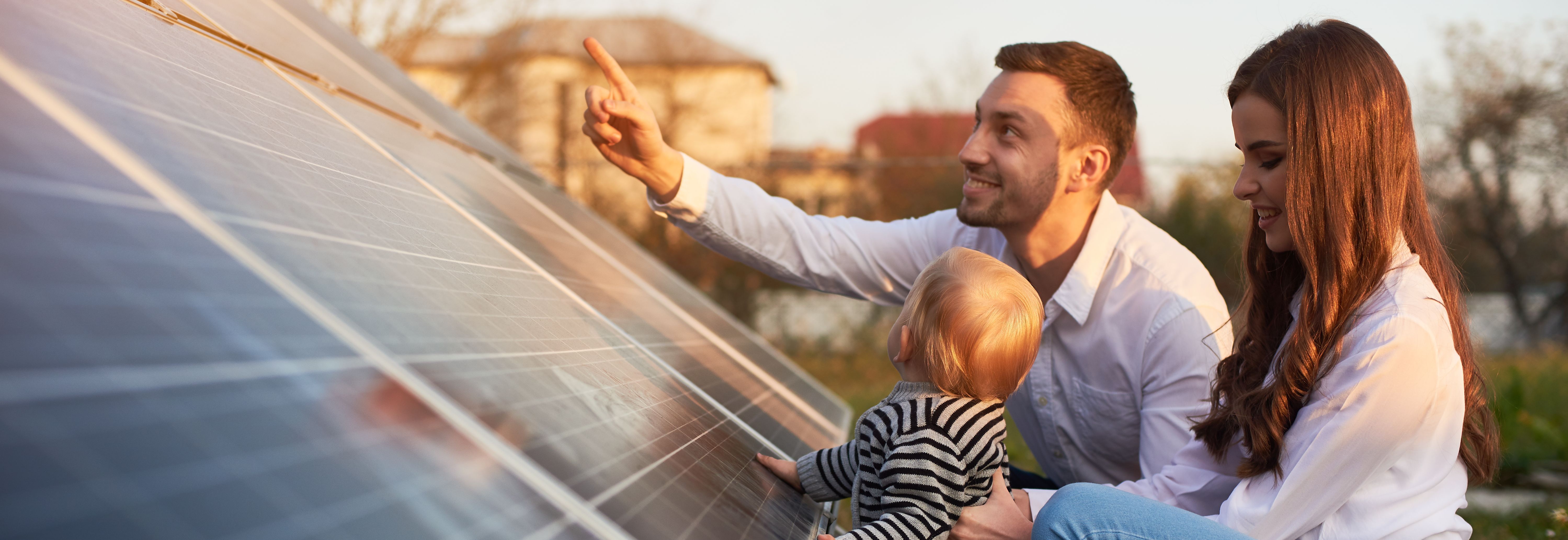 Ein Mann, eine Frau und ein kleines Kind betrachten gemeinsam eine Reihe von Solarpaneelen im Freien. Der Mann zeigt mit dem Finger auf etwas auf den Paneelen, während die Frau das Kind hält. Die Szene findet bei Tageslicht statt, und im Hintergrund sind Bäume und ein Gebäude zu sehen.