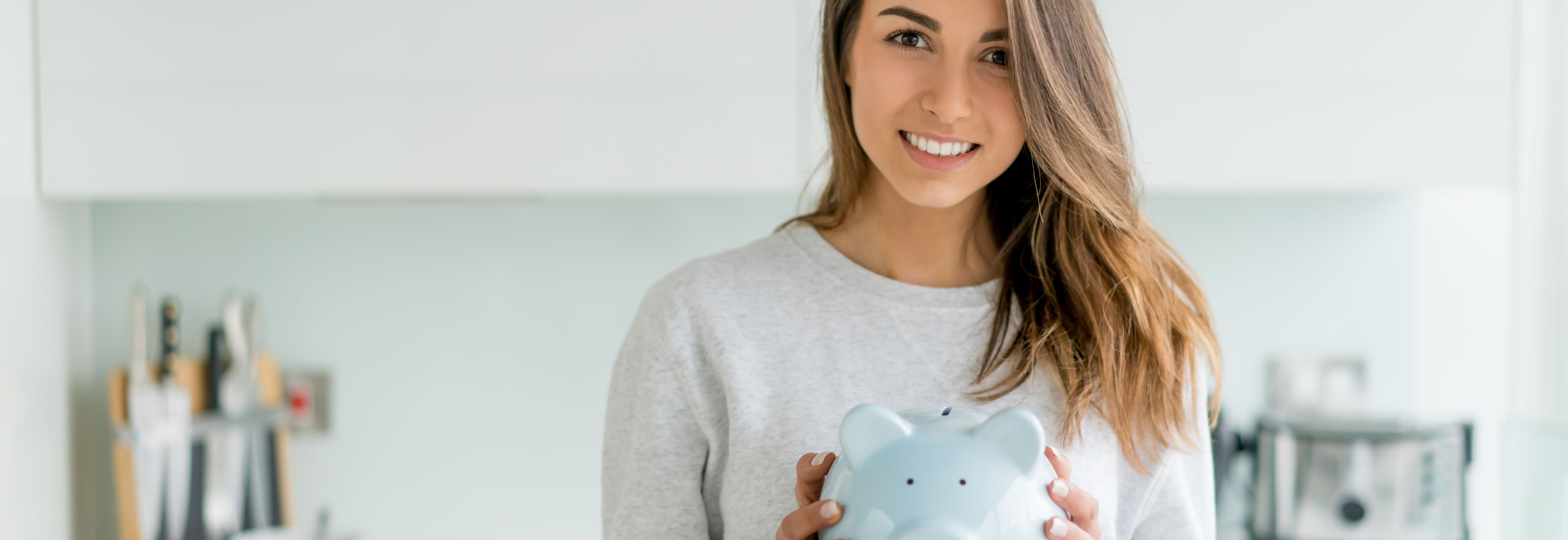 Eine Frau steht in einer Küche und hält ein blaues Sparschwein. Sie schaut geradeaus. Im Hintergrund sind Küchenutensilien, ein Toaster und andere Gegenstände auf der Arbeitsplatte zu sehen.