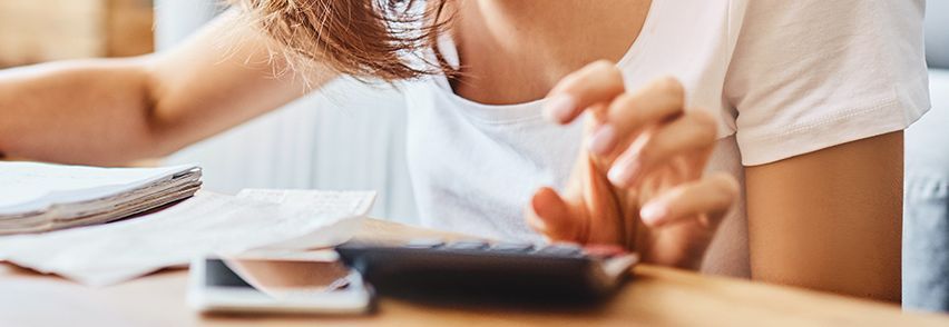 Eine Frau sitzt an einem Tisch und arbeitet mit einem Taschenrechner. Vor ihr liegen verschiedene Papiere und ein Smartphone. Im Hintergrund ist eine Uhr zu erkennen, die leicht verschwommen ist.