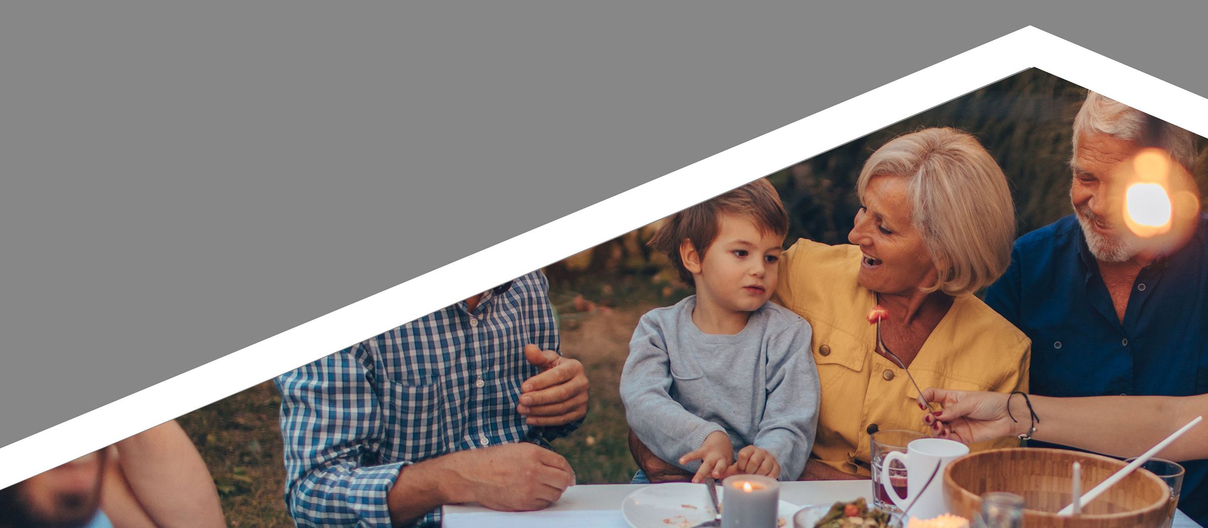 Mehrere Menschen sitzen draußen an einem Tisch und essen gemeinsam. Im Vordergrund sitzt ein Kind auf dem Schoß einer älteren Person, die freundlich mit dem Kind spricht. Links daneben ist eine weitere Person, deren Gesicht nicht ganz zu sehen ist. Rechts im Bild sitzen zwei andere Personen, die sich unterhalten. Im Hintergrund sieht man viel Grün – wahrscheinlich ein Garten oder Park. Oben links steht der Text ‚Bausparen für alle‘. Darunter befindet sich ein Button mit der Aufschrift ‚Mehr zu Bausparen für alle erfahren‘.