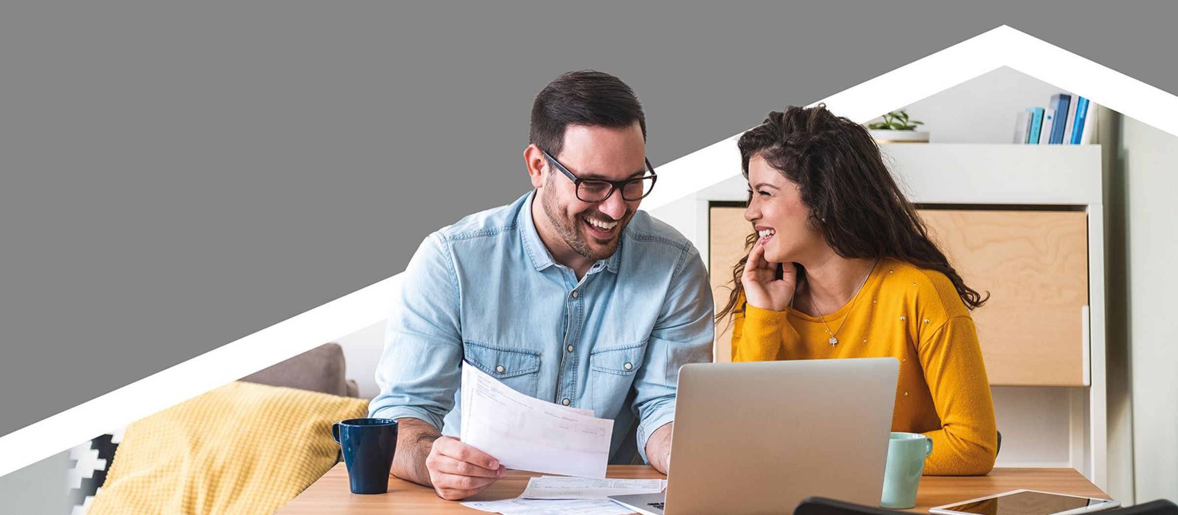 Ein Mann und eine Frau sitzen zusammen an einem Tisch. Der Mann trägt ein hellblaues Hemd und hält einige Papiere in der Hand, während die Frau, die ein gelbes Oberteil trägt, auf einen Laptop schaut. Im Hintergrund sind ein Sofa mit Kissen und ein Regal zu sehen.