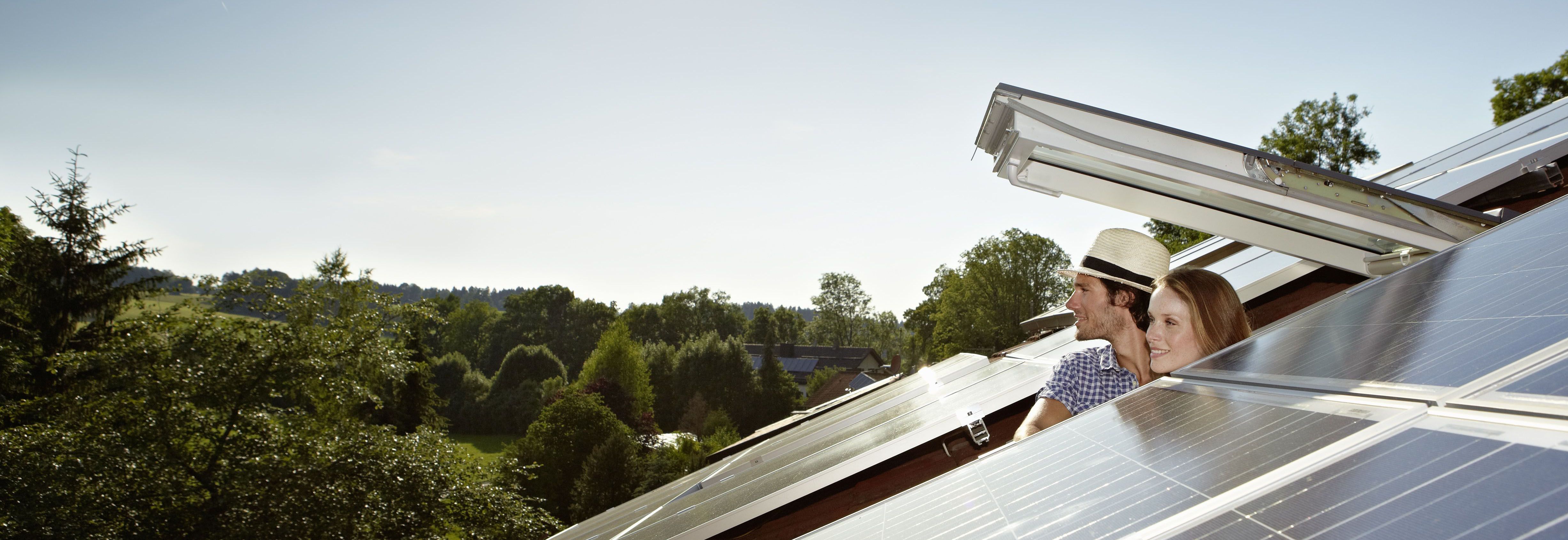 Das Bild zeigt zwei Personen, die auf einem Dach mit Solarpaneelen stehen. Eine Person trägt einen Strohhut und beide schauen in dieselbe Richtung. Die Solarpaneele sind glänzend und reflektieren das Sonnenlicht. Im Hintergrund sind Bäume und ein klarer Himmel zu sehen.