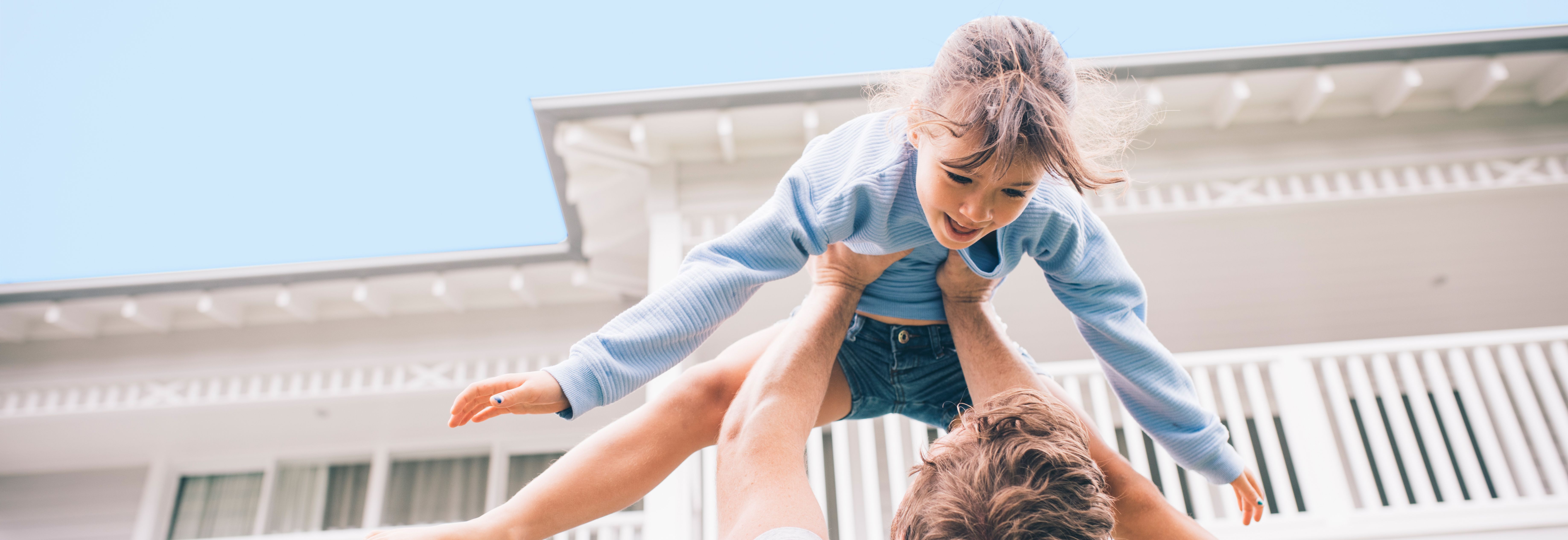 Ein Mann hebt ein Kind in die Luft, während sie beide lachen. Im Hintergrund ist ein weißes Haus mit einem Balkon und blauen Himmel zu sehen. Das Kind trägt einen blauen Pullover und eine kurze Hose.