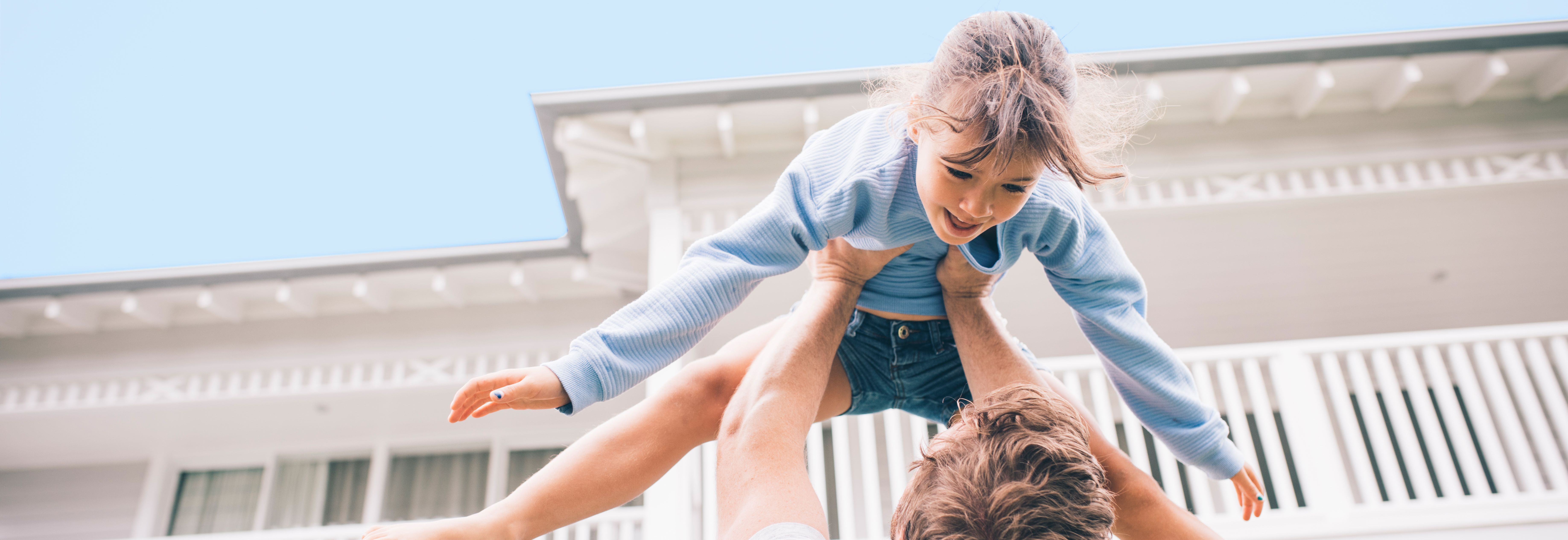 Ein Mann hebt ein Kind in die Luft, während sie beide lachen. Im Hintergrund ist ein weißes Haus mit einem Balkon und blauen Himmel zu sehen. Das Kind trägt einen blauen Pullover und eine kurze Hose.