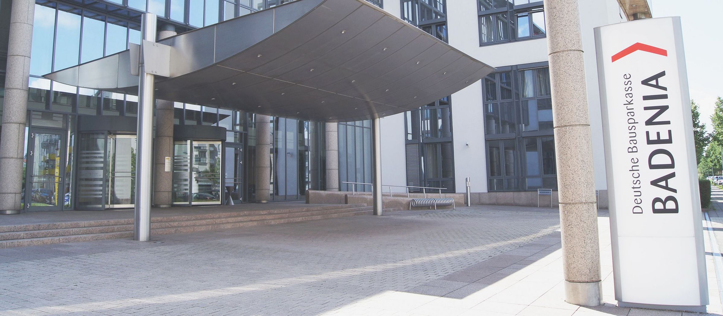 Das Bild zeigt den Eingangsbereich eines modernen Bürogebäudes. Links im Bild befindet sich eine Glasdrehtür, die in das Gebäude führt. Über dem Eingang erstreckt sich ein großes Vordach. Rechts im Vordergrund steht ein Schild mit der Aufschrift "Deutsche Bausparkasse BADENIA" und einem roten Dachsymbol. Das Gebäude hat große Fenster und eine helle Fassade. Vor dem Eingang sind Fahrradständer zu sehen, und der Boden ist mit Pflastersteinen bedeckt.