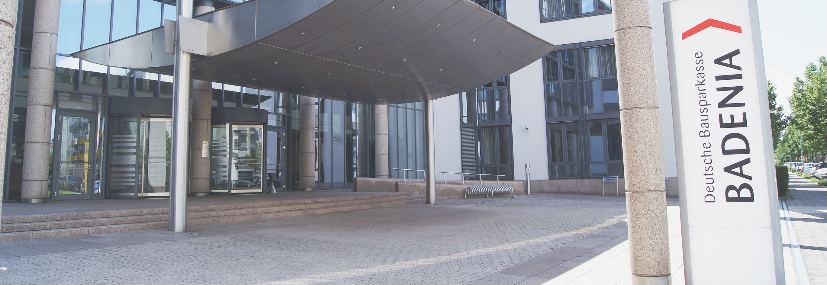 Das Bild zeigt den Eingangsbereich eines modernen Bürogebäudes. Links im Bild befindet sich eine Glasdrehtür, die in das Gebäude führt. Über dem Eingang erstreckt sich ein großes Vordach. Rechts im Vordergrund steht ein Schild mit der Aufschrift "Deutsche Bausparkasse BADENIA" und einem roten Dachsymbol. Das Gebäude hat große Fenster und eine helle Fassade. Vor dem Eingang sind Fahrradständer zu sehen, und der Boden ist mit Pflastersteinen bedeckt.
