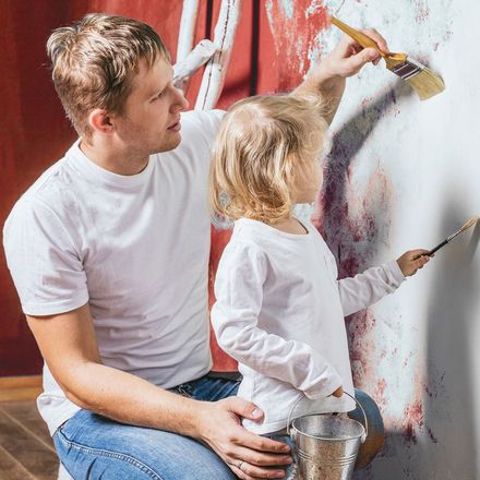 Ein Mann und ein Kind streichen gemeinsam eine Wand. Beide tragen weiße Oberteile und halten Pinsel in den Händen. Der Mann steht links und das Kind rechts, beide sind von hinten zu sehen. Die Wand hat bereits einige Farbflecken, während sie weiter daran arbeiten. Im Hintergrund ist eine Leiter zu erkennen.