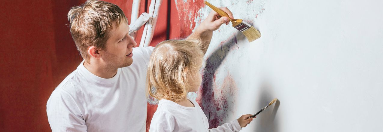 Ein Mann und ein Kind streichen gemeinsam eine Wand. Beide tragen weiße Oberteile und halten Pinsel in den Händen. Der Mann steht links und das Kind rechts, beide sind von hinten zu sehen. Die Wand hat bereits einige Farbflecken, während sie weiter daran arbeiten. Im Hintergrund ist eine Leiter zu erkennen.