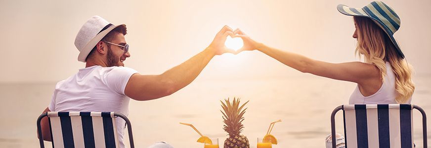 Das Bild zeigt ein Paar, das auf gestreiften Liegestühlen am Strand sitzt. Beide Personen tragen weiße Kleidung und Hüte. Sie halten ihre Hände zusammen in Form eines Herzens, durch das die Sonne scheint. Zwischen ihnen steht eine Ananas auf einem kleinen Tisch, daneben befinden sich zwei Getränke mit Strohhalmen.